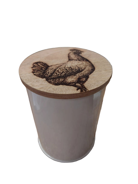 A white cylindrical container with a round wooden lid featuring a dark brown engraved bird design, shown on a wooden tabletop in a real indoor setting.